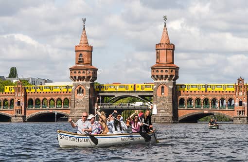backstagetourism-teamevent-06 Ruderboot mit dem Namen „Spritzmeister“ fährt vor der Oberbaumbrücke entlang, im Hintergrund fährt eine gelbe U-Bahn über die Brücke