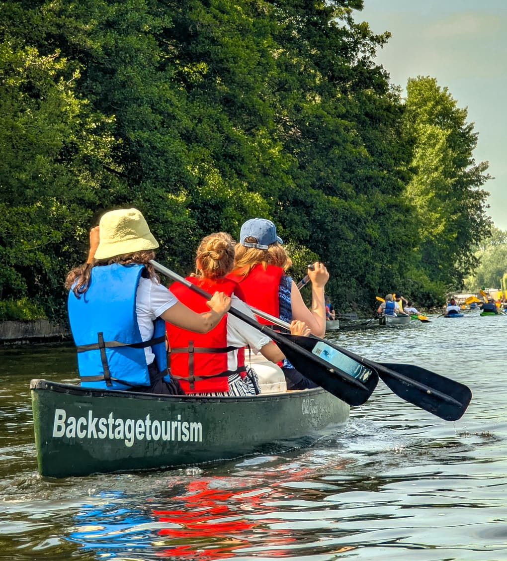 backstagetourism_tour-woc-02 Drei Personen mit Schwimmwesten paddeln in einem Backstagetourism-Kanu auf einem von Bäumen gesäumten Fluss